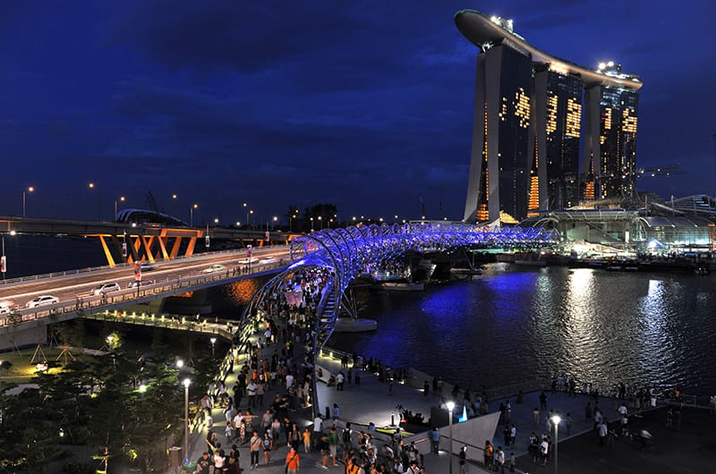 Night view of Singapore: Marina Bay Sands hotel, illuminated Helix Bridge with people, and waterfront promenade.